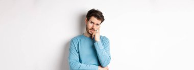 Kuşkucu genç adam sıkılmış yüzüyle seni dinliyor, kameraya gönülsüz bakıyor, beyaz arka planda eşofman üstü duruyor..