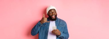 Mutlu siyah adam bana işaret veriyor, telefon ediyor, gülümsüyor, akıllı telefon tutuyor, bereyle ve kot gömlekle pembe arka planda dikiliyor..