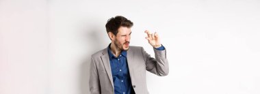 İş adamı küçük bir şey görmeye çalışıyor, parmaklarıyla küçük bir şey gösteriyor, beyaz arka planda takım elbise içinde duruyor..