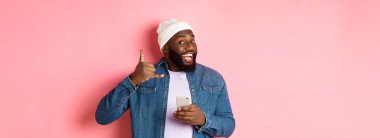 Mutlu siyah adam bana işaret veriyor, telefon ediyor, gülümsüyor, akıllı telefon tutuyor, bereyle ve kot gömlekle pembe arka planda dikiliyor..