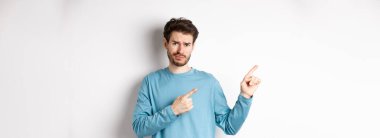 Şüpheci genç adam sakallı, tatminsiz, somurtkan, sol üst köşedeki logoyu işaret eden şüpheli, beyaz arka planda üzgün duruyor..