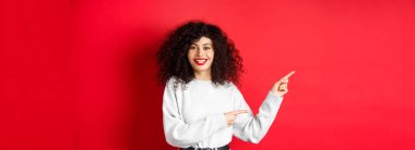 Kıvırcık saçlı, kırmızı dudaklı, gülümseyen beyaz kadın logoyu işaret ediyor, tanıtım teklifini gösteriyor, stüdyo geçmişine karşı duruyor..