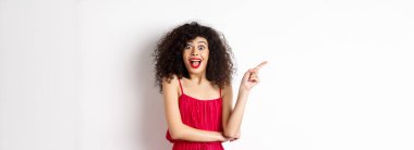 Heyecanlı, kıvırcık saçlı, makyajlı, zarif kırmızı elbiseli, şaşırmış ve beyaz arka planda duran logoyu işaret eden bir kadın..