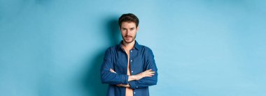 Kendine güveni tam genç bir adam kendini profesyonel hissediyor, kollarını göğsünde kavuşturuyor ve sırıtıyor, mavi arka planda duruyor..