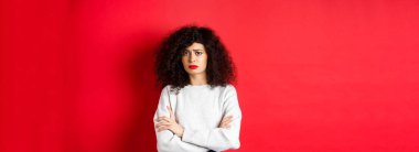 Üzgün ve endişeli beyaz kadın, kaşlarını çatmış göğsünde çapraz kollar ve endişeli görünüyor, kötü hissediyor, kırmızı arka planda duruyor..