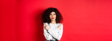 Düşünceli genç bir kadın seçim yapıyor, parmaklarını yana çeviriyor ve kendine güvenen yüzüyle sol üst köşeye bakıyor, kırmızı arka planda duruyor..