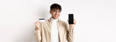 İnternetten alışveriş. Gözlüklü yakışıklı adam boş cep telefonu ekranı ve kredi kartı gösteriyor, gülümsüyor ve görüntüye bakıyor, beyaz arka planda duruyor..