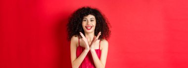 Kıvırcık saçlı, gece elbisesi giyen, el çırpan ve gülümseyen neşeli genç kadın, alkışlarla iyi performansı olan, kırmızı arka planda duran.