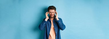 Hasta hisseden, başıyla temas eden, baş ağrısı çeken, baş dönmesi hisseden, mavi arka planda duran bir adam..