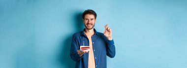 Kutlama ve tatil konsepti. Umut dolu genç adam doğum günü pastasında dilek diliyor, şans diliyor ve mavi arka planda durup olumlu gülümsüyor..