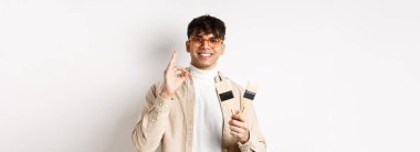 Yakışıklı, gözlüklü, boyama fırçalarını yenileme ve dekorasyon için gösteren, beyaz arka planda duran doğal bir adam..