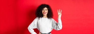 Üç numarayı gösteren, gülümseyen, sipariş veren, kırmızı arka planda eşofman üstü giyen neşeli bir kadın model..