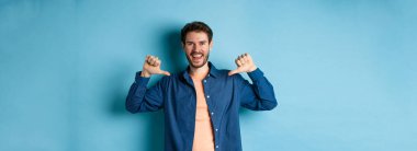Kendi reklamını yapan mutlu genç adam, kendini işaret ediyor ve gülümsüyor, mavi arka planda profesyonel gibi duruyor..