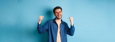 Neşeli genç adam kutlama yapıyor, elini kaldırıyor ve evet diyor, gözlük giyiyor, mavi arka planda duruyor..