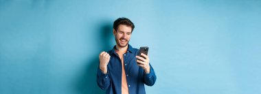 Şanslı adamın çevrimiçi kazandığı, cep telefonu ekranını okuduğu ve yumruk pompası yaptığı, zafer kazandığı, mavi arka planda durduğu görüntü.