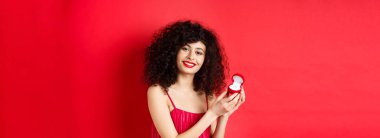 Kıvırcık saçlı, kırmızı elbiseli ve nişan yüzüğünü gösteren romantik bir kadın stüdyo arka planında mutlu duruyor..