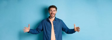 Mutlu gülümseyen adam ellerini yana açıp büyük bir nesne gösteriyor. Büyük nesnelerden bahsediyor. Mavi arka planda neşeli duruyor..