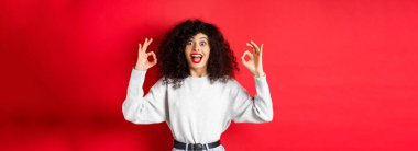 Heyecanlı kız iyi işaretler gösterir, mutlu gülümser, iyi bir şey önerir, övgüyü hak eder, kırmızı arka planda şaşkına döner..