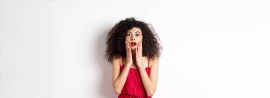 Heyecanlı kıvırcık saçlı kadın, inançsızlıkla yüz yüze, kameraya şaşkın şaşkın bakıyor, tanıtıma bakıyor, beyaz arka planda kırmızı bir kadın gibi duruyor..