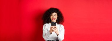 Gülümseyen modern kız akıllı telefondan fotoğraf çekiyor, ekrana bakıyor ve video kaydediyor, kırmızı arka planda duruyor..