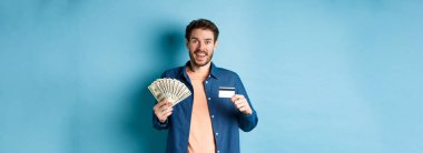 Gülümseyen beyaz adam plastik kredi kartı ve para gösteriyor, mutlu görünüyor, mavi arka planda duruyor..