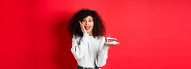 Heyecanlı bir kadın doğum gününü kutluyor, elinde pasta tutuyor ve kameraya şaşırmış ve mutlu bakıyor, kırmızı arka plan.