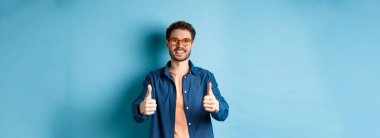 Gözlüklü mutlu genç adam başparmağını kaldırıyor, gözlükçü dükkanını tavsiye ediyor, mavi arka planda duruyor..