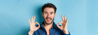 Sakallı adamın imzasını atıp gülümsemesi, harika bir seçimi övmesi, harika bir şeyi övmesi, mavi arka planda dikilmesi..