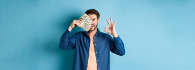 Neşeli genç adam tamam işareti gösteriyor ve yüzünün yarısını dolarlarla kaplıyor, mavi arka planda duruyor..