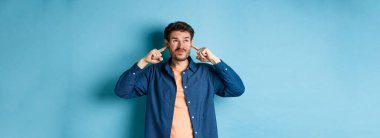 Sinirli genç adam sesi, parmaklarıyla kulaklarını kapatıyor ve yüzünü buruşturuyor, yüksek sesli müzikten rahatsız oluyor, mavi arka planda duruyor..
