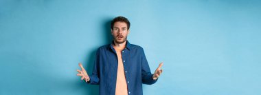 Şok olmuş genç adam ellerini kaldırıyor ve kameraya bakıyor. Korkmuş, inançsızlıkla bakıyor, mavi arka planda duruyor..