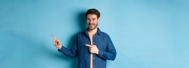 Sakallı neşeli beyaz adam boş bir yeri işaret ediyor ve kameraya gülümsüyor. Reklam yapıyor, mavi arka plan..