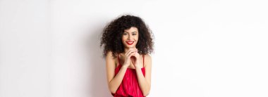 Kurnaz, kıvırcık saçlı, çan kulesi gibi parmakları olan, sinsi gülümseyen, bir planı olan, beyaz arka planda kırmızı elbiseli genç bir kadın..