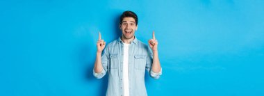 Mutlu 25 'li yaşlarda sakallı, parmaklarını kaldırıp gülümseyen, reklamını yapan, mavi arka planda duran bir adamın portresi..