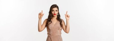 Yeni yıl, Noel ve kutlama konsepti. Kırmızı rujlu zarif bir genç kadın, parti elbisesi giyiyor ve küstah görünüyor, logoyu işaret ediyor, beyaz arkaplan.