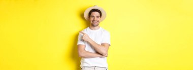 Genç erkek turist sağı işaret ediyor, gülümsüyor ve reklam veriyor, turizm ve yaşam tarzı kavramı, sarı arka plan.