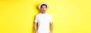 Hasır şapkalı şaşkın adam mutlu görünüyor, seyahat acentası reklamına hayret ediyor, sarı arka planda duruyor..