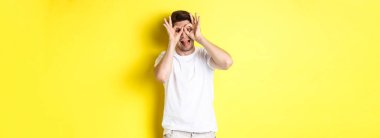 Genç adam komik yüzler ve yapışkan dil gösteriyor, sarı arka planda şakacı duruyor..