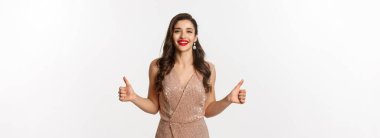 Kutlama, tatil ve parti kavramı. Lüks elbiseli, başparmaklarını kaldıran ve gülümseyen, onaylayan, beyaz arka planda duran,.