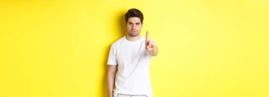 Hayır diyen kendine güvenen bir adam, bir şeyi reddeder ve yasaklar, bir parmağını gösterir ve kaşlarını çatar, sarı arka planda durur..