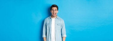 Gözlüklü çekici, mutlu adam şaşırmış görünüyor, reklamlara bakıyor, mavi arka planda duruyor..