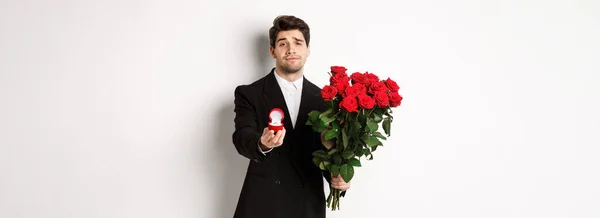 stock image Handsome smiling man in black suit, holding roses and engagement ring, making a proposal to marry him, standing against white background.