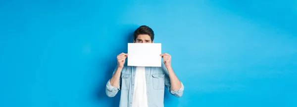Shy and nervous guy hiding face behind blank piece of paper, peeking with worried look, showing your logo, standing over blue background.