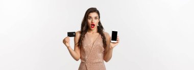 İnternetten alışveriş ve tatil konsepti. Kadın şaşırmış görünüyor ve beyaz arka planda duran akıllı telefon ekranlı kredi kartı gösteriyor..