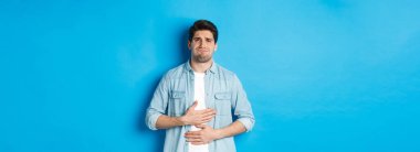 Karnının üstünde el ele tutuşan ve acıdan suratını buruşturan, karın ağrısı çeken, mavi arka planda duran bir adam..