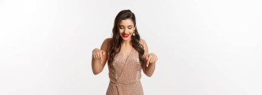 Kırmızı rujlu güzel bir kadın, Noel partisi için şık bir elbise giyiyor, parmaklarını yere eğip gülümsüyor, beyaz arka planda dikiliyor..