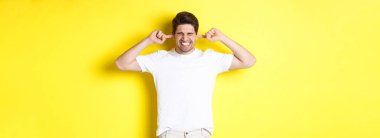 Sinirli adam suratını buruşturup kulaklarını kapatıyor, gürültüden şikayet ediyor, sarı arka planda duruyor. Boşluğu kopyala