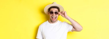 Turizm ve tatil kavramı. Yazlık şapkalı ve güneş gözlüklü mutlu adamın yakın çekimi. Tatilin tadını çıkarıyor, sarı arka planda duruyor..