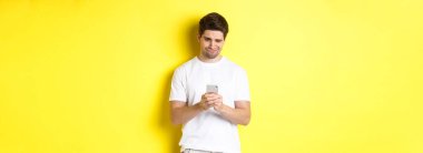 Akıllı telefon ekranından rahatsız olmuş gibi görünen, telefonda garip mesajlar okuyan, beyaz tişörtlü, sarı arka planda duran bir adam..