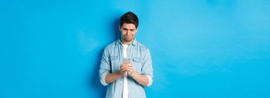 Hayal kırıklığına uğramış ve şüpheci adam telefona bakıyor, mavi arka planda duruyor..